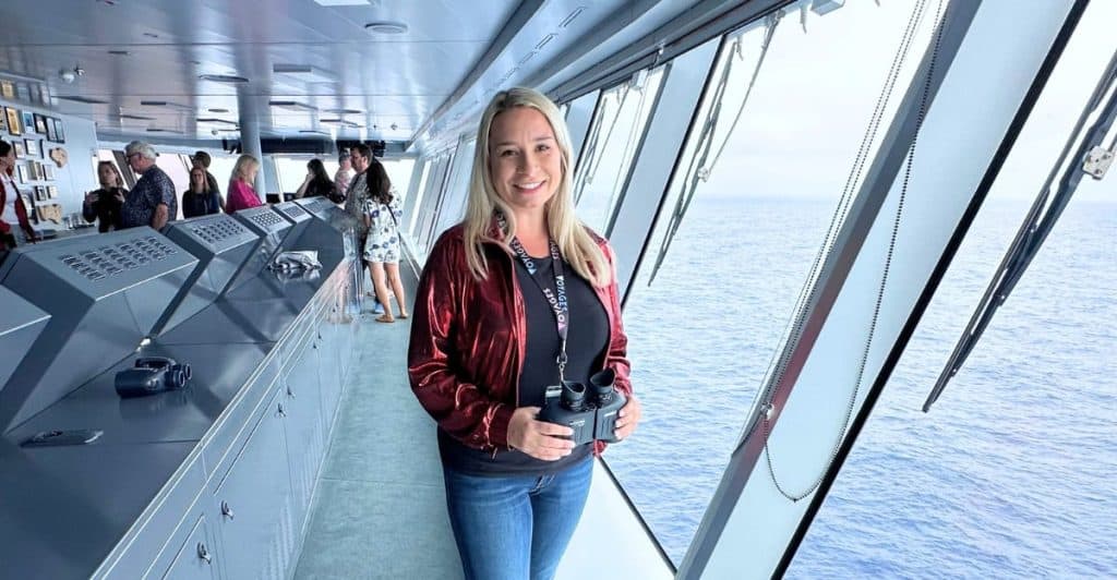 Woman holding binoculars while touring the bridge of a Virgin Voyages ship with large windows overlooking the ocean. The behind the scenes bridge visit is part of her Virgin Voyages Mega RockStar cruise experience.