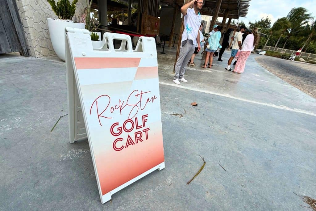 A sidewalk sign reading "RockStar Golf Cart" outside a resort entrance where guests wait for transportation.