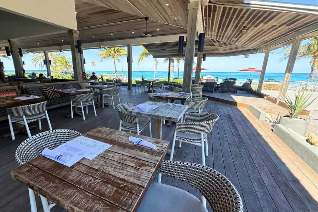 Richard's Retreat access for Rockstar and MegaRockstar Virgin Voyages sailors. Open air beachfront restaurant with weathered wood tables and woven chairs overlooking palm trees and bright turquoise ocean.