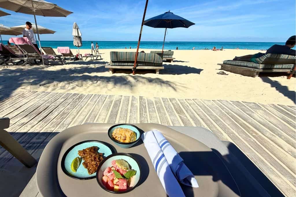 Service at Richard's Retreat for Rockstar and MegaRockstar sailors. Small plates of food served at a shaded beachfront table with lounge chairs and umbrellas stretching toward the turquoise ocean.