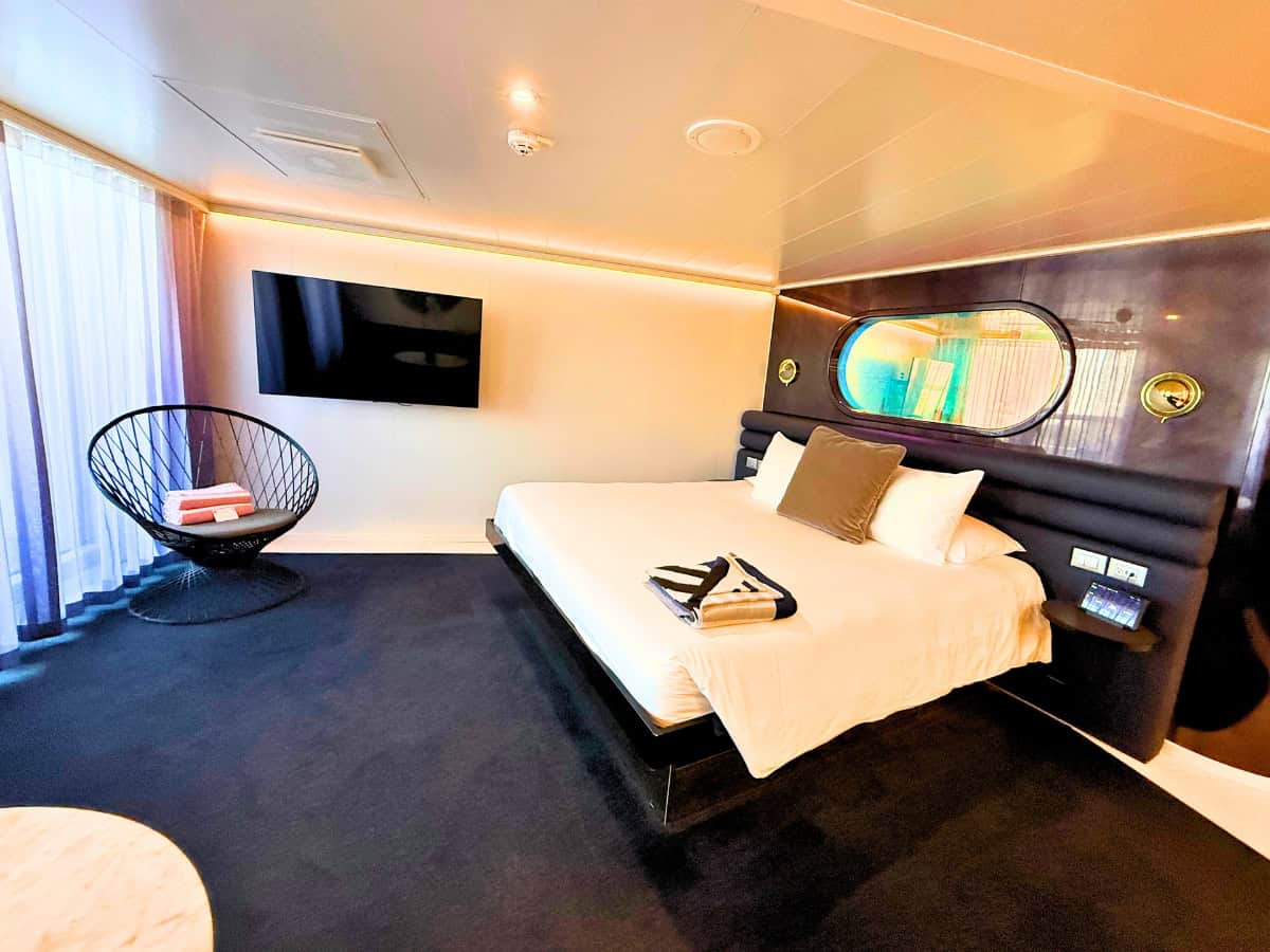Bedroom inside a Virgin Voyages Mega RockStar Gorgeous Suite with a large white bed. A round window above the headboard and warm accent lighting give the room a modern nautical feel.