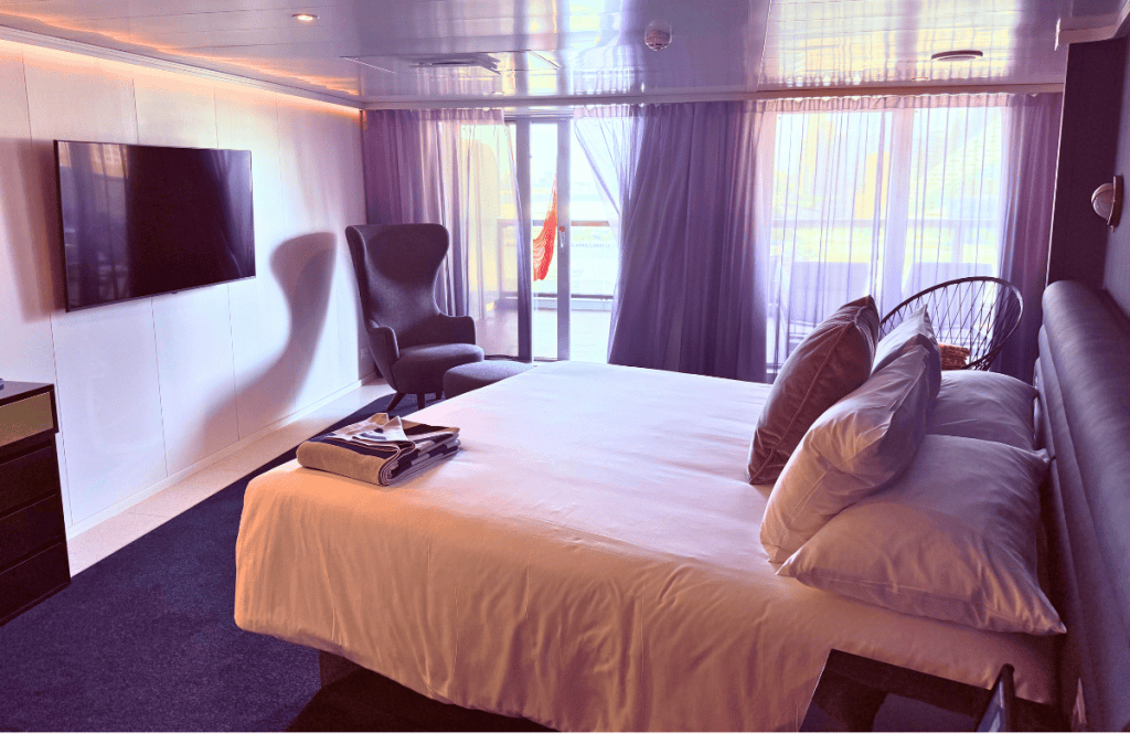 Virgin Voyages Mega RockStar bedroom with a large white bed wall mounted television and seating area. Floor to ceiling glass doors open onto the private terrace.