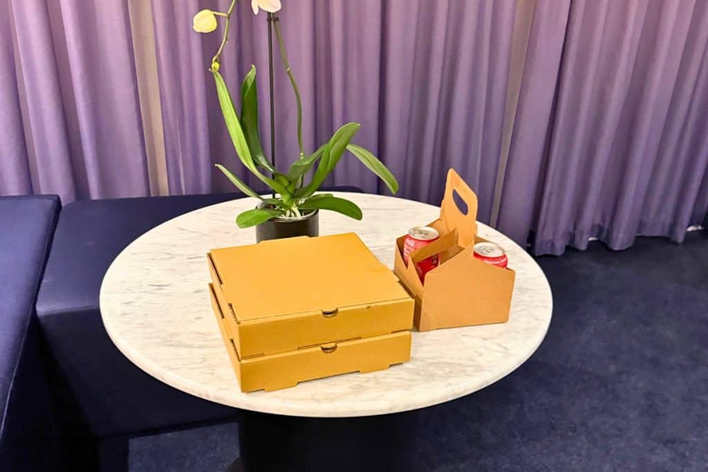 In-cabin food delivery for Virgin Voyages MegaRockstars. Two stacked pizza boxes and a drink carrier with two cans arranged beside an orchid on a marble table inside the suite.