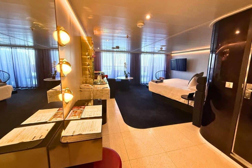 Spacious Virgin Voyages Mega RockStar suite with a large bed lounge area stocked bar vanity and floor to ceiling windows leading to the balcony.