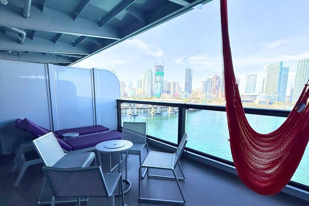 MegaRockstar Gorgeous Suite private cruise ship balcony with lounge chairs a small table and a red hammock overlooking blue water and the Miami skyline.