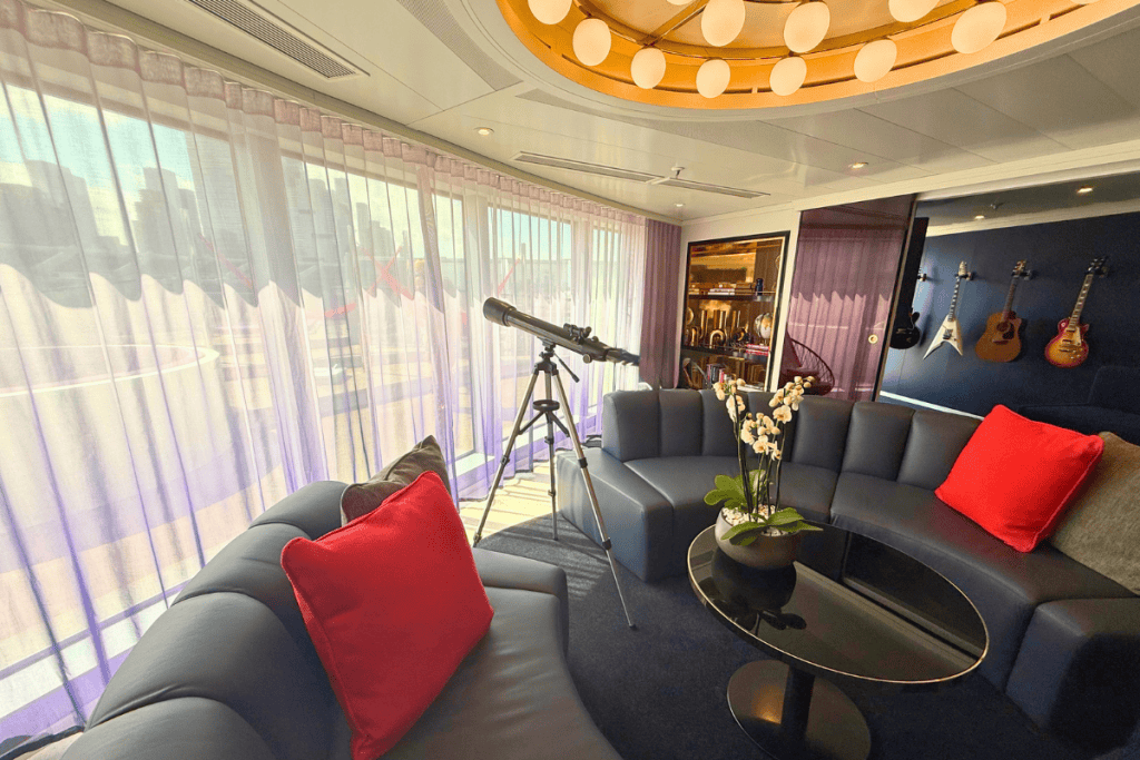 Spacious suite lounge with curved dark sofas red pillows a telescope and guitars displayed on the wall. Floor to ceiling windows overlook the private terrace.