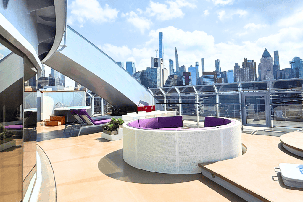 Expansive private cruise ship terrace with circular purple seating loungers and a hot tub overlooking the Manhattan skyline. The outdoor space is part of a Virgin Voyages Mega RockStar suite.