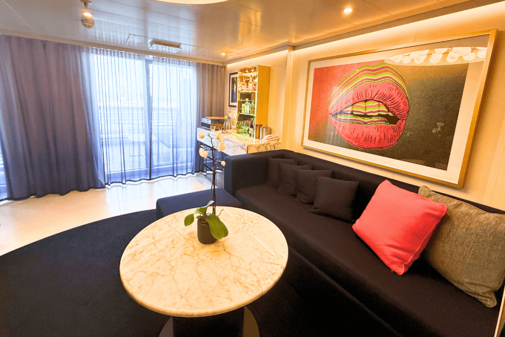 Virgin Voyages Mega RockStar suite living area with a dark sofa marble table orchid and colorful artwork of lips. A stocked bar sits beside glass doors leading to the terrace.