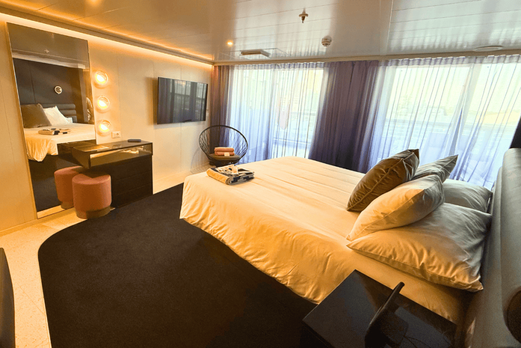 Large bedroom with a white bed vanity television and floor to ceiling windows leading to the private terrace. Warm lighting and dark furnishings give the Virgin Voyages Mega RockStar suite a modern feel.