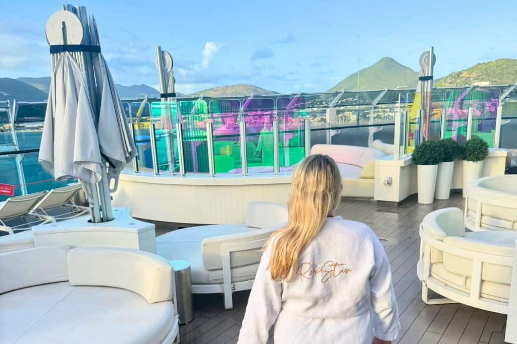 Eleanor wearing a white robe embroidered with "RockStar" while walking across Richard's Rooftop outdoor lounge deck. White seating colorful glass panels and green Caribbean mountains surround the Virgin Voyages Mega RockStar guest experience.