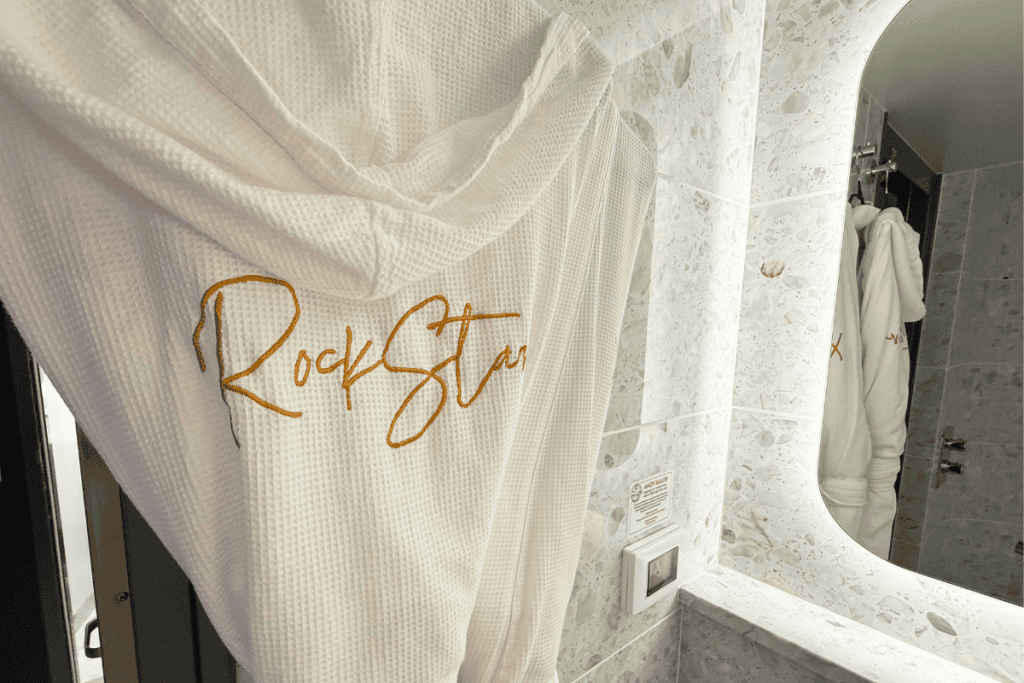 White waffle robe embroidered with "RockStar" hanging inside a marble bathroom. A second matching robe is reflected in the illuminated mirror.