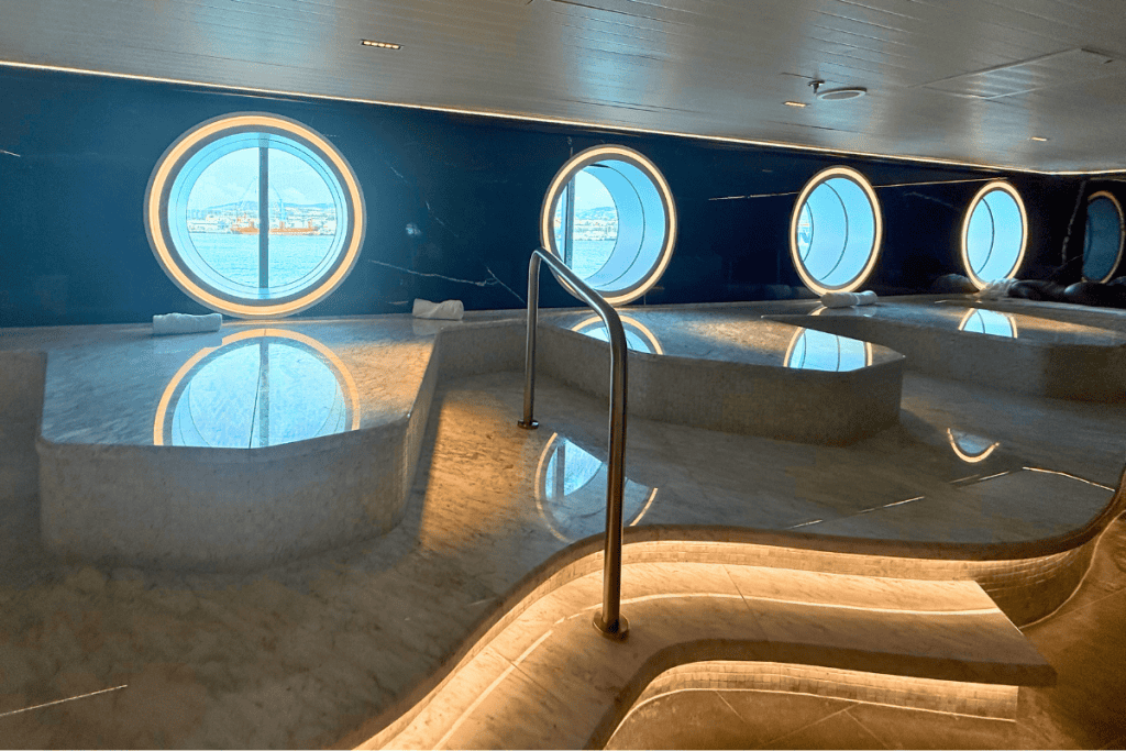 Marble Redemption Spa room aboard Virgin Voyages with heated stone loungers and curved illuminated steps. Round porthole windows overlook the water while the polished surfaces reflect the natural light.